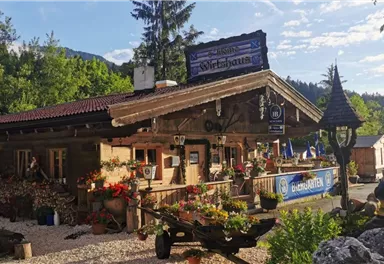 Ein gemütliches Holzhaus im bayerischen Stil, umgeben von Blumen und grüner Natur. Die Fassade ist einladend dekoriert und hat einen rustikalen Charme.