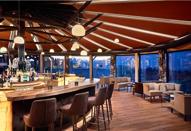 A modern bar with a large circular counter and stylish seating. Large windows offer a view of the surroundings at dusk.