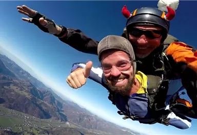 Ein mutiger Tandemsprung mit einem scherzenden Fallschirmspringer. Im Hintergrund sind die beeindruckenden Berglandschaften zu sehen.