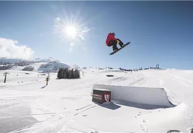 A snowboarder jumps over a kicker in the snow. The sky is clear and the sun is shining.