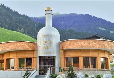 Ein modernes Gebäude in Form einer Flasche befindet sich in einer malerischen Berglandschaft. Grüne Wiesen und Hügel umgeben die Architektur.