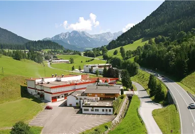An idyllic landscape with green hills and mountains in the background. In the foreground, several buildings and a highway can be seen.