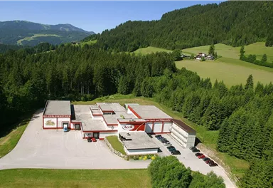 A modern building amidst a green landscape with mountains in the background. Pavilions and parking lots are clearly visible.