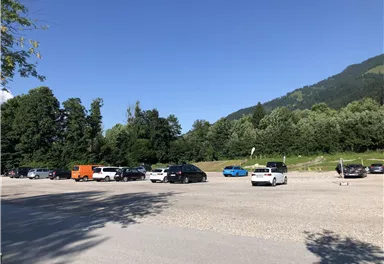 Ein großer Parkplatz, der teilweise gefüllt ist, umgeben von Bäumen und Bergen. Der Himmel ist klar und blau.
