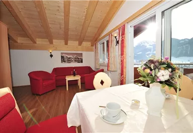Ein gemütliches Wohnzimmer mit rotem Sofa und Tisch. Große Fenster bieten einen Blick auf die Berge und viel Tageslicht.