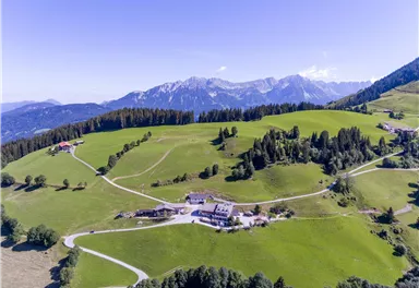 Eine grüne Berglandschaft mit sanften Hügeln und vereinzelten Hütten. Im Hintergrund sind hohe Berge und ein klarer blauer Himmel zu sehen.