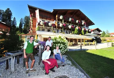 Ein traditionelles Café im bayerischen Stil mit blühenden Blumen und einem gepflegten Garten. Vier Personen in Trachten stehen vor dem Gebäude und lächeln freundlich.