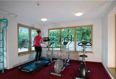 Ein modernes Fitnessstudio mit verschiedenen Geräten. Eine Person trainiert auf einem Ellipsentrainer vor großen Fenstern mit Blick ins Grüne.