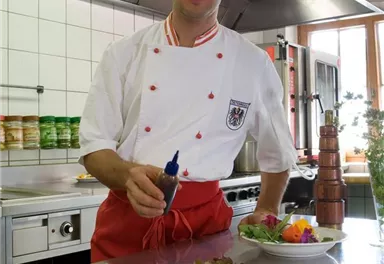 Ein Koch steht in einer modernen Küche und präsentiert einen kunstvoll angerichteten Teller mit Salat und Gemüse. Im Hintergrund sind Küchengeräte und weitere Gerichte sichtbar.
