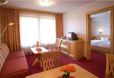 Ein gemütlicher Wohnbereich mit einem Sofa, einem Tisch und einem Fernseher. Im Hintergrund ist ein Schlafzimmer mit einem Doppelbett sichtbar.