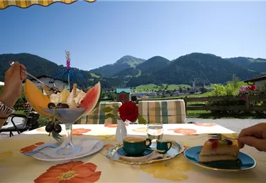 Ein gedeckter Tisch mit einer großen Schale Obstsalat und einem Stück Kuchen. Im Hintergrund sind grüne Berge und ein blauer Himmel zu sehen.
