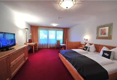 Ein gemütliches Hotelzimmer mit einem großen Doppelbett und einem Fernseher. Der Raum ist hell und freundlich, mit einem Tisch und Stühlen sowie einem schönen Ausblick durch das Fenster.
