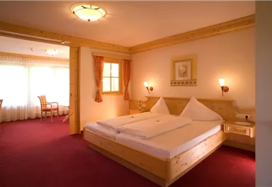 Ein gemütliches Hotelzimmer mit einem großen Bett und Holzdetails. Es gibt einen sonnigen Sitzbereich mit einem Fenster und gemütlicher Atmosphäre.