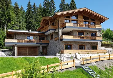 Ein modernes Haus aus Holz und Stein, umgeben von grünen Bäumen und Rasen. Der klare Himmel und die ruhige Umgebung schaffen eine einladende Atmosphäre.