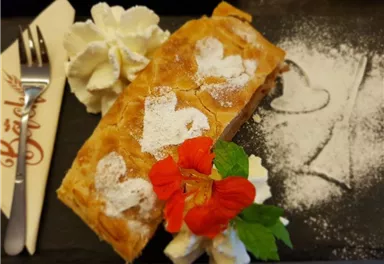 A delicious apple strudel with powdered sugar and decorative hearts made of dough. Served with cream and fresh flowers on an elegant background.