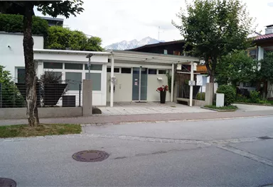 A modern building with large windows and an entrance area. In the foreground, trees and a quiet street are visible.