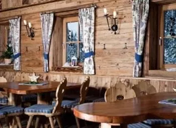 Ein gemütlicher Innenraum mit Holzwänden und großen Fenstern. Die Tische sind aus Holz und mit blauen Sitzkissen ausgestattet.