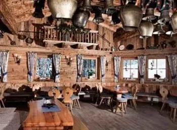 Ein gemütlicher, holzverkleideter Raum mit rustikalen Tischen und Stühlen. An der Decke hängen verschiedene Glocken, und durch die Fenster ist die winterliche Landschaft sichtbar.