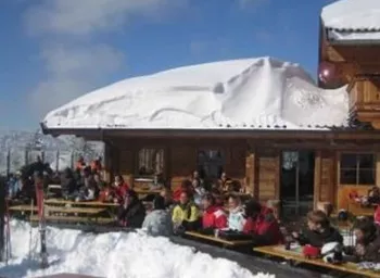 Eine Skihütte mit vielen Gästen im Freien, umgeben von Schnee. Der Himmel ist blau und klar.