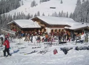 Ein belebtes Skihüttengelände im Schnee mit vielen Skifahrern und Skiausrüstungen. Im Hintergrund sind verschneite Bäume und Berge zu sehen.