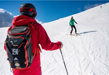 A skier in red clothing stands on a snow-covered slope. In the background, another person in green clothing is going down the run.
