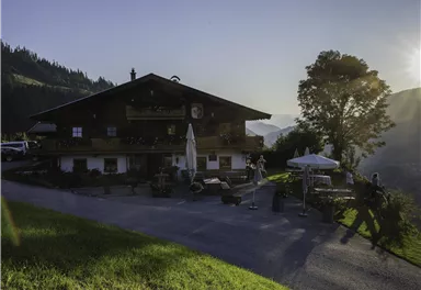 A traditional mountain house surrounded by nature. The sun rises behind the mountains and illuminates the landscape.