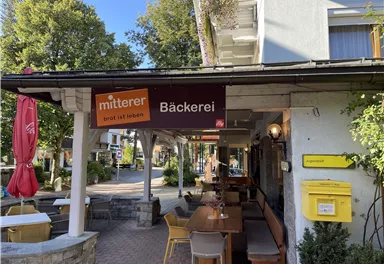 A cozy bakery with an inviting outdoor area. The surroundings are characterized by trees and sunny weather.