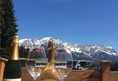 A bottle of champagne stands next to glasses on a table with a view of snow-covered mountains. The clear blue sky highlights the pleasant atmosphere.