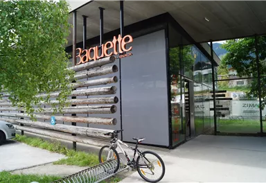 A modern shop with the sign "Baguette". Outside, there is a bicycle and a green area with trees.