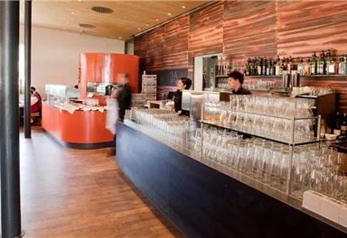 Ein modernes Restaurantinnere mit einem langen Bartresen und vielen Gläsern. Im Hintergrund sieht man Mitarbeiter, die Gäste bedienen.