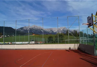 Ein Basketballplatz mit roten Belag in den Bergen. Im Hintergrund sind grüne Wiesen und schneebedeckte Gipfel zu sehen.