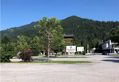 A tranquil landscape with green trees and a view of the mountains. In the foreground, there is a square with unpaved ground and buildings in the background.