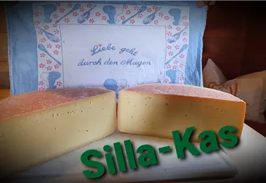 Zwei große Käselaibe stehen auf einem weißen Teller. Im Hintergrund liegt ein dekoratives Tuch mit der Aufschrift „Liebe geht durch den Magen“.