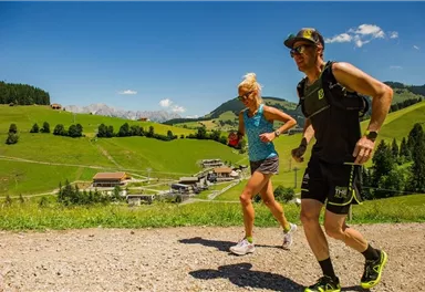 Two people are jogging on a gravel path in a picturesque green landscape. In the background, hills and small houses are visible.