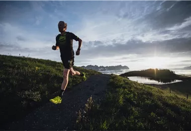 A runner jogs on a path in a picturesque landscape. In the background, mountains and a lake can be seen at sunrise.
