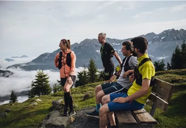 A group of four athletes enjoys the view of the mountains. They are standing and sitting on a bench, surrounded by green scenery and fog.