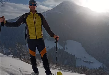 A person is standing in the snow with snowshoes and a mountain in the background. The landscape is wintry and sunny.