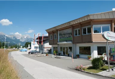 A modern building with large windows stands on a street. In the background, mountains and a clear blue sky are visible.