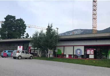 A modern building with colorful facades and a parking lot in the foreground. In the background, there are trees and a green hill visible.