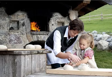 A woman and a small girl are kneading dough in front of a traditional wood-fired oven. In the background, a green meadow and stones are visible.