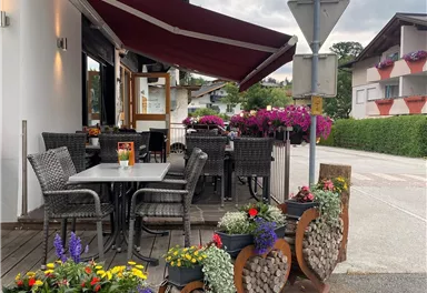 Ein gemütlicher Außenbereich eines Restaurants mit Tischen und Stühlen. Bunte Blumenampeln und dekorative Pflanzen verleihen dem Platz eine einladende Atmosphäre.
