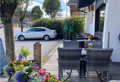 Ein einladender Außenbereich mit Rattanmöbeln und bunten Blumenarrangements. Im Hintergrund parkt ein Auto vor einem gepflegten Gebäude.