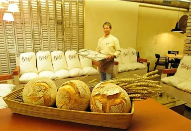 A baker presents fresh bread in a cozy café. In the foreground, there are decoratively arranged loaves and decorative ears of grain.