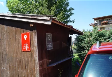 A wooden cabin with a red electric car next to it. There are signs for a Tesla charging station and general information.