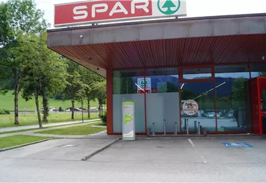 A SPAR supermarket with large windows and a modern entrance area. In the background, trees and a green meadow can be seen.
