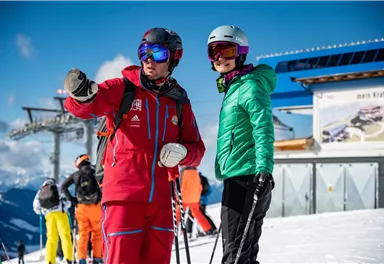A ski instructor is giving a student instructions on the slope. In the background, other skiers and a ski lift can be seen.
