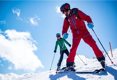 Two skiers are skiing down the slope. The sky is blue with a few clouds.