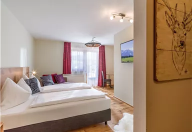 Ein gemütliches Zimmer mit zwei Einzelbetten, hellen Farben und einem Fernseher. Große Fenster mit Vorhängen bieten einen Blick nach draußen.