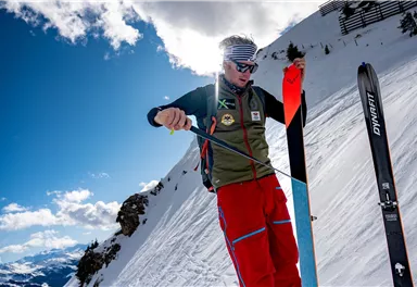A skier on a snow-covered slope is preparing his equipment. In the background, there are mountains and a blue sky.
