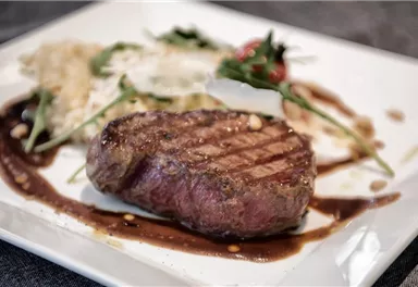 A juicy piece of beef on a white plate, served with fresh salad and a sauce. In the background, there is a slightly creamy side dish.